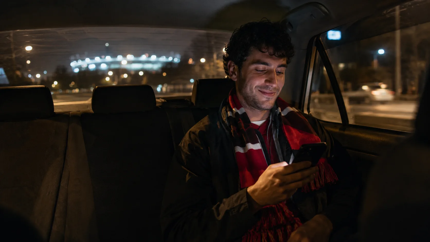 Soccer fan in a red and white striped scarf relaxing in the back seat of a rideshare car at night with stadium lights glowing in the distance